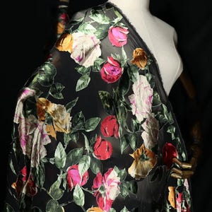 May include: A sheer black fabric featuring a vibrant floral pattern of roses in shades of red, pink, yellow, and white, interspersed with green leaves. The textile is draped over a mannequin, showcasing the detailed velvet-like floral texture.