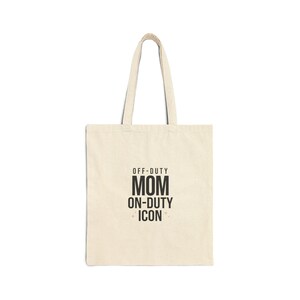 May include: Cream-colored tote bag with black text that reads "OFF-DUTY MOM ON-DUTY ICON". The bag has two handles and is made of a canvas-like material. The text is centered on the front of the bag.