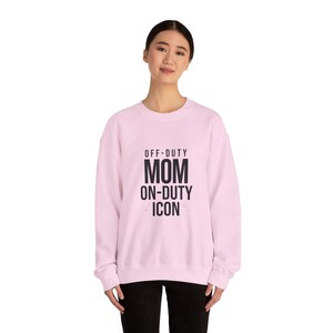 May include: A light pink sweatshirt with the text "OFF-DUTY MOM ON-DUTY ICON" in black. The sweatshirt is a crew neck style and appears to be made of a soft material. The overall theme is casual wear.