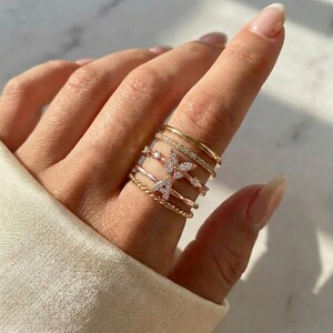 Peut inclure: Une main porte plusieurs bagues dorées aux designs variés. On distingue une bague papillon, une bague torsadée et des anneaux ornés de détails scintillants. Les bagues, de tailles et de styles différents, créent un effet superposé.