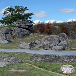 Puede incluir: Vista exterior con grandes rocas grises, hierba verde y una carretera. Una persona está sentada en las rocas. El cielo es azul con nubes. Un árbol está encima de las rocas. Escena natural y pintoresca.