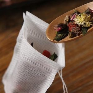 May include: A white, rectangular, non-woven fabric tea bag with a heat-sealed edge. A wooden spoon is positioned above the open top of the tea bag, containing a mixture of dried yellow chrysanthemum flowers, red goji berries, and green leaves.