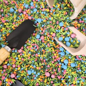 May include: A vibrant sensory bin filled with colourful sprinkles in various shapes and sizes, including blue, pink, yellow, and orange spheres and star shapes. Two black garden trowels and two wooden scoops are also present.