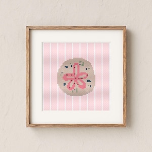 May include: A framed cross-stitch art piece featuring a pink sand dollar design on a tan background. The background has vertical pink and white stripes. The artwork is displayed in a wooden frame, suitable for coastal or beach-themed decor.