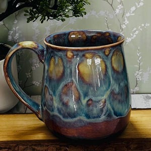 May include: A ceramic mug featuring a glossy glaze with a blend of deep blue, teal, and warm amber tones. The surface displays a mottled, reactive pattern with darker brown accents near the base and a curved handle on the left side.