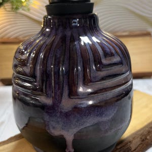K&ouml;nnte beinhalten: Ein Keramik-Seifenspender mit abgerundetem K&ouml;rper, der eine tiefviolette und schwarze Glasur mit gl&auml;nzendem Finish aufweist. Die Oberfl&auml;che hat ein erhabenes geometrisches Muster mit Tropfen aus hellerer violetter Glasur und einen mattschwarzen Pumpkopf.