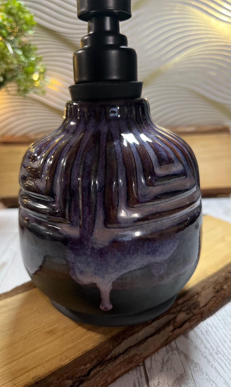 K&ouml;nnte beinhalten: Ein Keramik-Seifenspender mit abgerundetem K&ouml;rper, der eine tiefviolette und schwarze Glasur mit gl&auml;nzendem Finish aufweist. Die Oberfl&auml;che hat ein erhabenes geometrisches Muster mit Tropfen aus hellerer violetter Glasur und einen mattschwarzen Pumpkopf.