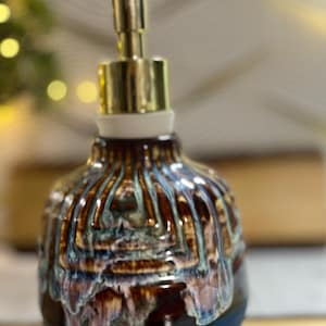 May include: A ceramic soap dispenser with a glossy, multi-colored glaze featuring vertical drips of brown, cream, and blue over a solid dark teal base. The dispenser is topped with a metallic gold-toned pump mechanism and a white collar.
