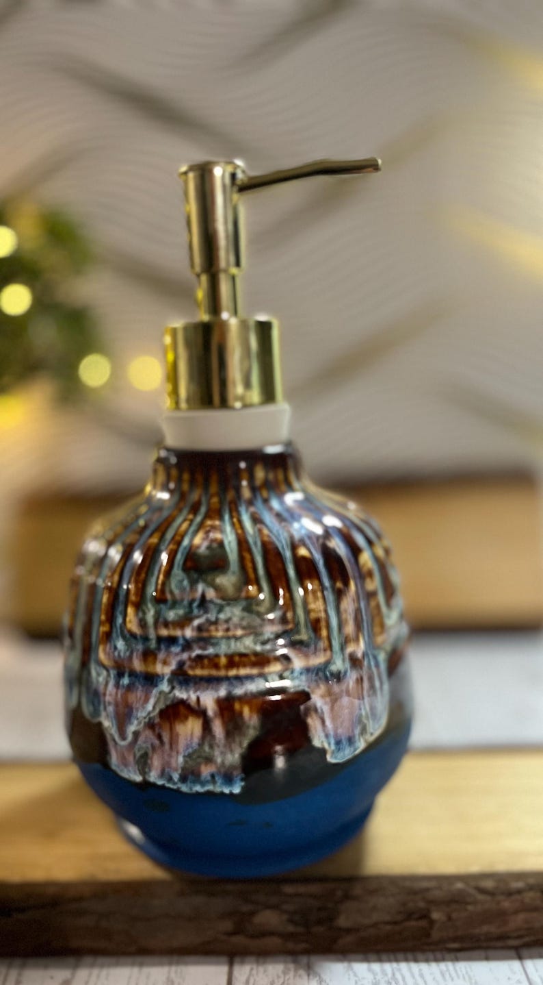 May include: A ceramic soap dispenser with a glossy, multi-colored glaze featuring vertical drips of brown, cream, and blue over a solid dark teal base. The dispenser is topped with a metallic gold-toned pump mechanism and a white collar.