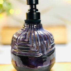 K&ouml;nnte beinhalten: Ein Keramik-Seifenspender mit bauchigem K&ouml;rper, der eine tiefviolette und schwarze Glasur mit einem texturierten geometrischen Muster aufweist. Der Spender ist mit einem mattschwarzen Kunststoffpumpmechanismus versehen, der horizontal nach rechts ragt.