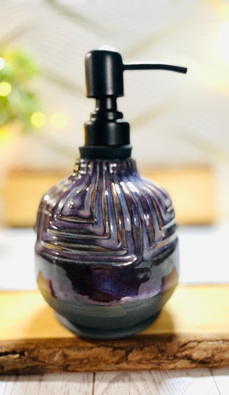 K&ouml;nnte beinhalten: Ein Keramik-Seifenspender mit bauchigem K&ouml;rper, der eine tiefviolette und schwarze Glasur mit einem texturierten geometrischen Muster aufweist. Der Spender ist mit einem mattschwarzen Kunststoffpumpmechanismus versehen, der horizontal nach rechts ragt.