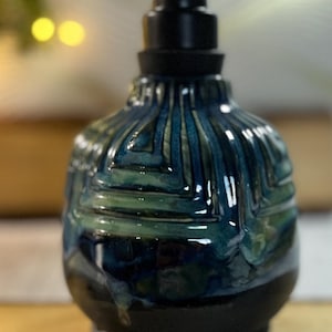 May include: A ceramic soap dispenser with a bulbous body featuring a glossy, textured glaze in shades of deep blue and forest green. The base is a solid matte black, and the top is fitted with a functional black plastic pump mechanism.