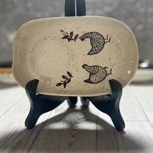 May include: An oval ceramic grater plate with a speckled cream finish. The surface features two illustrated chickens and two small leaf sprigs in black and red. A circular textured grating area is on the left, and three small holes are on the right.