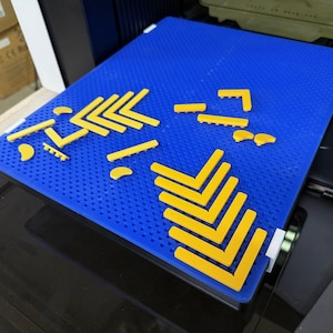 Könnte beinhalten: Eine blaue 3D-Drucker-Bauplatte mit einem Raster aus kleinen Löchern, auf der mehrere gelbe Kunststoffkomponenten angeordnet sind. Die gelben Teile umfassen mehrere V-förmige Eckwinkel, gerade Stangen und kleine gebogene Segmente.
