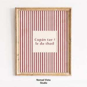 May include: A framed wall art print featuring a vertical striped pattern in dark red and cream. In the centre, a cream-coloured rectangular label with a scalloped edge displays the text: Cupán tae ? le do thoil. The frame is light brown wood.