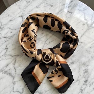 May include: A tan and black leopard print scarf with a gold-coloured heart-shaped clasp adorned with small, clear crystals. The scarf has black borders and is displayed on a marble surface.