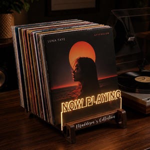 May include: A wooden vinyl record display stand featuring an illuminated acrylic sign that reads "NOW PLAYING" in a bold font. The base includes the engraved text "Maddison's Collection." A row of vinyl records is held upright in the stand.