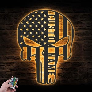 May include: A custom-made skull-shaped wall art piece with an American flag design. The left side features stars, while the right side has stripes. The text "CUSTOM NAME" is vertically displayed. The piece is illuminated with a warm glow.