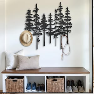 May include: A black metal wall art piece depicting a forest of pine trees. A straw hat and keys hang from the trees. A dog leash hangs from the right side. Below, a bench with pillows, baskets, and shoes.