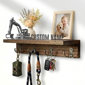Peut inclure: Une étagère murale en bois foncé avec quatre crochets en laiton antique. L'étagère présente une silhouette d'excavatrice en métal noir, un vase avec des fleurs séchées et un cadre photo en bois clair avec un enfant tenant un chiot.
