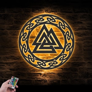 May include: A circular black wall art piece featuring a central Valknut symbol enclosed within an intricate Celtic knotwork border. The design is backlit with a warm orange glow, creating a high-contrast silhouette against the wall surface.
