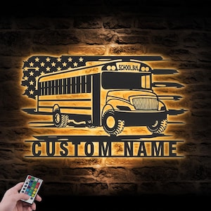 Può includere: Un cartello di scuolabus in metallo nero con un design a bandiera americana e le parole "SCHOOL BUS" in alto. Il cartello ha le parole "CUSTOM NAME" sotto l'autobus. Il cartello è illuminato da un bagliore caldo contro un muro di mattoni.