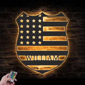 Può includere: Decorazione murale a forma di scudo con il design della bandiera americana e il nome "WILLIAM" illuminato in oro. Le stelle e le strisce sono visibili, con una trama simile a mattoni sullo sfondo. Un telecomando è nell'angolo in basso a sinistra.