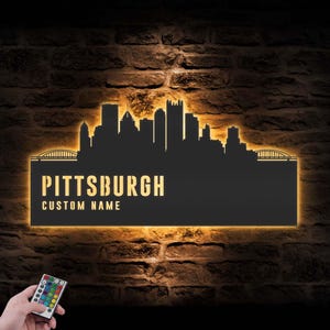 Peut inclure: Panneau métallique noir représentant la silhouette de l'horizon de Pittsburgh, avec le nom de la ville et un espace pour un nom personnalisé. Le panneau est éclairé d'une lueur chaude sur un mur de briques. Une télécommande avec des boutons colorés est en bas à gauche.