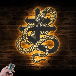 May include: A black metal wall art piece featuring a snake coiled around a double-barred cross, illuminated by a warm orange LED backlight. The design includes a small handheld remote control with multiple colored buttons for light adjustment.