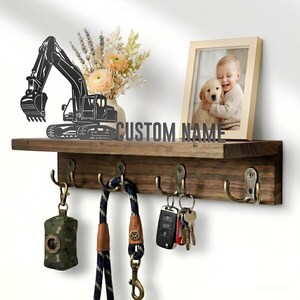 Peut inclure: Une étagère murale en bois foncé avec quatre crochets en laiton antique supportant un distributeur de sacs à déjections, une laisse et des clés. L'étagère présente une silhouette d'excavatrice noire, le texte CUSTOM NAME, un vase et un cadre.