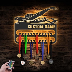 Puede incluir: Expositor de medallas de metal negro con temática de natación con el texto "CUSTOM NAME". La pantalla presenta una silueta de nadador y soportes para medallas con cintas de colores. Un mando a distancia está en la esquina inferior izquierda.