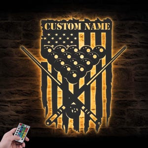 May include: A black metal wall art piece featuring a design of a pool table with crossed cues, a rack of billiard balls, and an American flag motif. The words "CUSTOM NAME" are at the top. The piece is illuminated with a warm, golden light.