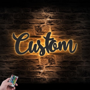 May include: A black "Custom" sign with a warm yellow glow against a textured brick wall. A hand holds a remote control with colorful buttons, suggesting customizable lighting options for the sign.
