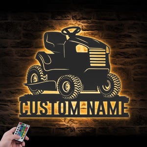 May include: A black metal wall sign shaped like a riding lawn mower with a backlit LED glow. The sign features a horizontal base plate with the text CUSTOM NAME in bold capital letters. A small remote control with multiple colour buttons is included.