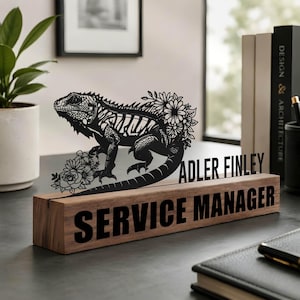 Op de afbeelding: Een houten bureau-naamplaatje met een zwarte metalen uitsparing van een leguaan omringd door bloemen. De naam "ADLER FINLEY" staat in zwarte letters boven de voet, met de titel "SERVICE MANAGER" gedrukt op de voorzijde van het houten blok.
