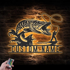May include: Black metal wall art showing a fish leaping from water, with a fisherman and fishing line. The words "CUSTOM NAME" are below the fish. A remote control with colour options is in the lower left.