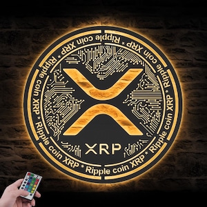 Puede incluir: Un letrero redondo e iluminado con el logotipo de la criptomoneda XRP. El letrero tiene un esquema de color dorado y negro, con el símbolo XRP en el centro. El texto "Ripple coin XRP" está escrito alrededor del borde. Un mando a distancia está en la esquina inferior izquierda.