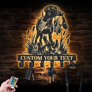 May include: A black metal wall-mounted key holder featuring a silhouette of a hunting dog carrying a duck amidst cattails, illuminated by a warm orange backlight. The base includes six hooks and the text CUSTOM YOUR TEXT with a remote control.
