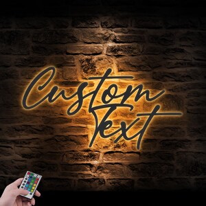 May include: A custom text sign with a warm yellow glow against a dark brick wall. The words "Custom Text" are written in a stylish script font. A hand holds a remote control with colorful buttons.