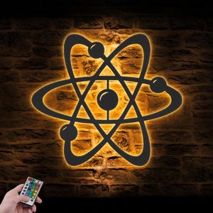 May include: A black atom-shaped wall light fixture featuring a central nucleus and three orbiting electron paths. The design is backlit with a warm orange glow, creating a silhouette effect. A small remote control with multiple color buttons is included.
