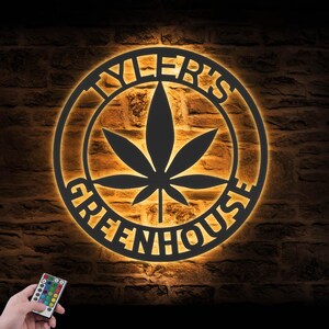 May include: A black metal sign with the words "TYLER'S GREENHOUSE" in a circular design, featuring a cannabis leaf in the centre. The sign is illuminated with a warm, orange glow, and a remote control is visible in the lower left corner.