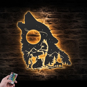 May include: A black metal wall art piece shaped like a howling wolf silhouette, backlit with warm orange LED lighting. The cutout design features a moon, a smaller wolf, pine trees, and mountain peaks. A small remote control is held in the foreground.
