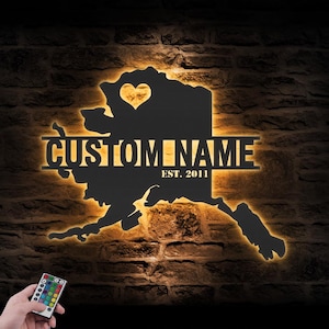 May include: Black metal wall art in the shape of Alaska, with a heart cutout and the words "CUSTOM NAME" and "EST. 2011". The art is illuminated with warm light, and a remote control is visible.