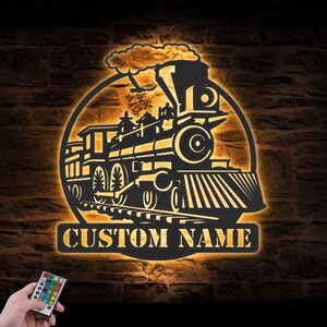 May include: A black metal train silhouette with a glowing yellow outline, set against a brick wall. The design includes a steam engine, tracks, and the text "CUSTOM NAME" in a rectangular banner. A remote control is in the lower left.