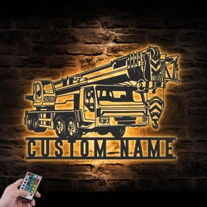 May include: A backlit metal sign featuring a crane truck silhouette. The sign is illuminated with a warm glow against a brick wall. The text "CUSTOM NAME" is displayed below the truck. A remote control is visible in the lower left corner.