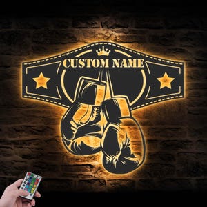 May include: A black and gold boxing-themed wall art piece with the text "CUSTOM NAME" above a pair of boxing gloves. The art features a crown and stars, and is illuminated with a warm glow. A remote control is visible in the lower left corner.
