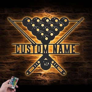 May include: A black metal wall art piece featuring a billiards theme. The design includes a rack of billiard balls, crossed cues, and the text "CUSTOM NAME" in illuminated lettering. The background is a brick wall.