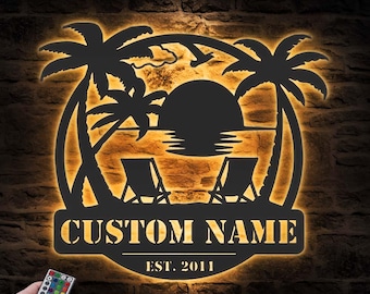 Puede incluir: Una pieza de arte mural de metal negro con una escena de playa tropical, con palmeras, una puesta de sol y dos tumbonas. Se muestran las palabras "CUSTOM NAME" y "EST. 2011". El arte está iluminado con un cálido resplandor naranja.
