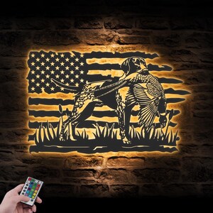 May include: A metal wall art piece depicting a hunting dog with a pheasant against an American flag backdrop. The artwork is illuminated with a warm glow, creating a striking silhouette. The dog is in a hunting stance, holding the pheasant in its mouth.