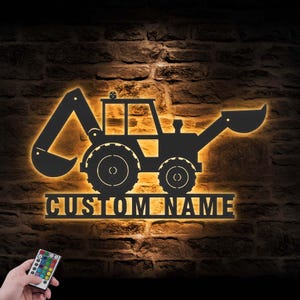 May include: A black metal excavator silhouette with the words "CUSTOM NAME" illuminated in a warm glow. The image is set against a textured brick wall, with a remote control in the lower left corner.